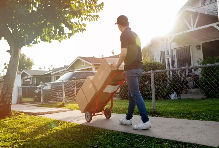 A person pushing a hand truck with several boxes in a residential neighborhood. The person is walking along the sidewalk in front of a house, indicating that they are likely delivering or picking up packages.